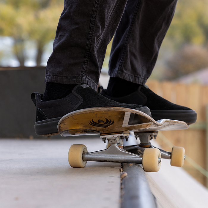 A close-up action shot of a person skateboarding on a concrete ledge, wearing black slip-on REV SOS Carlsbad barefoot shoes and dark jeans, with a worn skateboard in front.